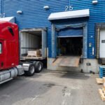 Camion semi-remorque rouge stationné à un quai de chargement d'entrepôt logistique avec nivellement hydraulique et porte sectionnelle